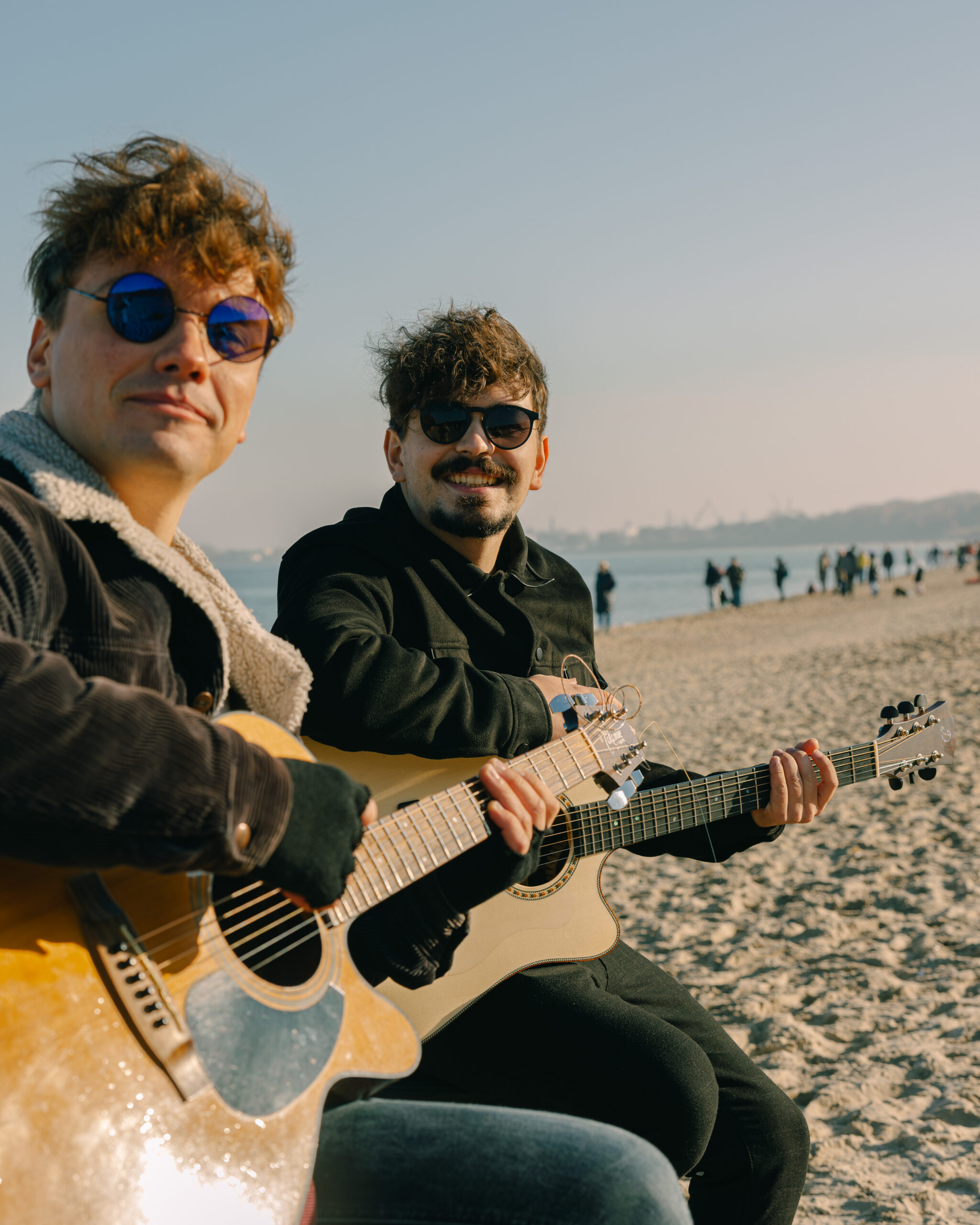 Zdjęcie dwóch członków zespołu AlterCollective – Kuby i Emila – grających na gitarach akustycznych na plaży. Obaj noszą okulary przeciwsłoneczne i ciepłe ubrania. W tle widać morze i spacerujących ludzi. Zdjęcie oddaje relaksacyjną i artystyczną atmosferę.